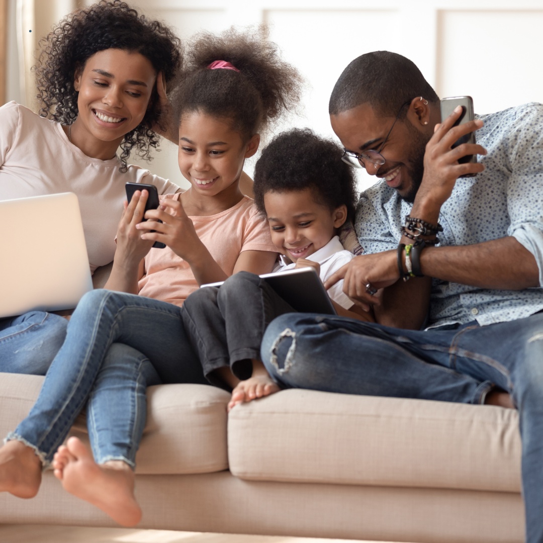 Happy Black family of four sitting together on a couch, parents and children enjoying quality time with technology devices, representing the warm community connections fostered at Blue Quill Development